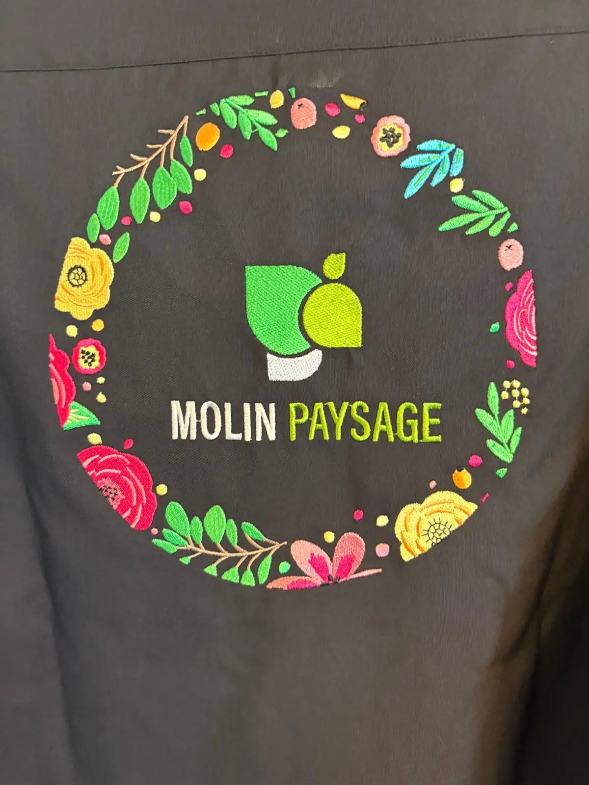 Logo Molin Paysage entouré d'une couronne de fleurs brodée en couleur dans le dos d'une veste noire